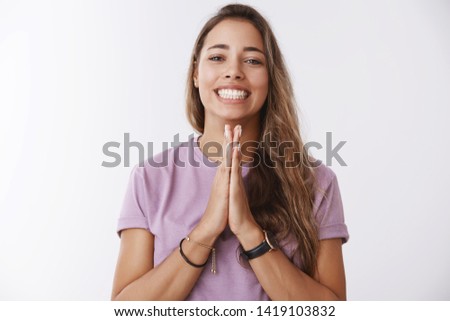 Clingy female friend asking favour smiling broadly holding hands pray grinning begging help advice pressing palms together under jawline, hopefully gazing camera, standing white background
