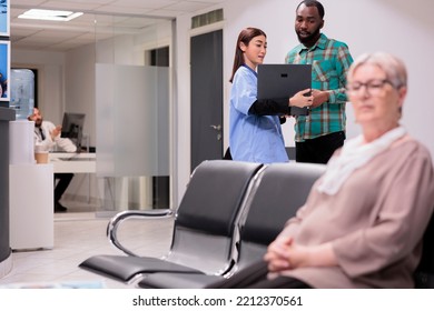 Clinic Emergency Medical Department Physician In Charge Searching For Young African American Male Electronic Records On Laptop. Elderly Caucasian Woman Sitting Waiting Hospital Admission.