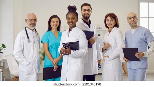 Clinic or hospital staff at work. Diverse team of happy smiling doctors, clinicians, therapists, cardiologists in scrubs and white coat uniform standing together, holding clipboards, looking at camera