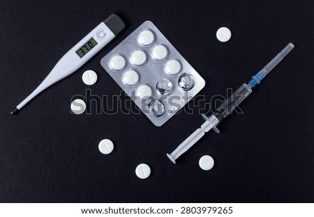 Clinical flat lay with digital thermometer showing 37.0, pills, blister pack and syringe on black background. Healthcare and medical treatment concept