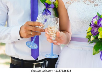 Clinking wedding glasses with champagne close-up by bride and groom in their hands on the backdrop of a white wedding dress, shirt and tie, bouquet and boutonniere of purple and yellow eustoma flowers