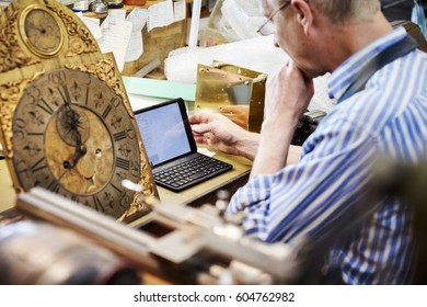 A clock maker in his workshop using a laptop Antique clock face - Powered by Shutterstock