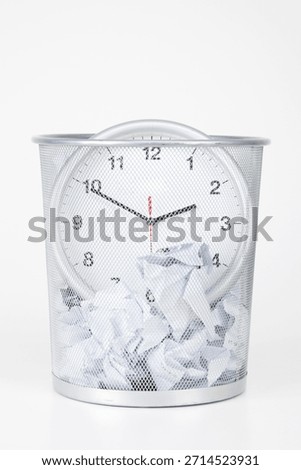 Clock placed inside a wire trash bin filled with crumpled paper at a work desk