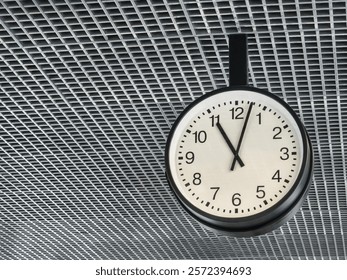 The clock shows exactly twelve o'clock in a contemporary setting. The design features clear numbers on a white face, suspended from a grid-patterned ceiling.