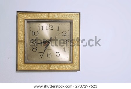 The clock is square in shape. It features a decorative outer frame consisting of four golden, textured panels that have a 