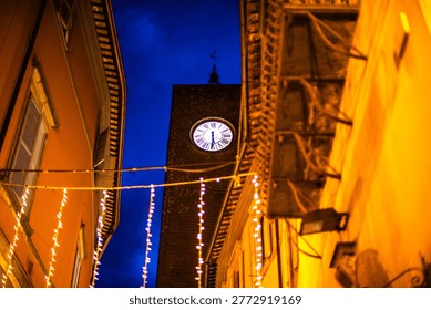 Clock tower rising between narrow buildings with decorative lights at night. Urban architecture scene suitable for travel, tourism, and historic city environment concepts.