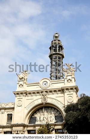 The clock tower stands atop the Edificio de Correos y Telégrafos (Post and Telegraph Building) in Valencia. Visitors look at the architecture while enjoying the sunny weather. The scene captures local life in the city.