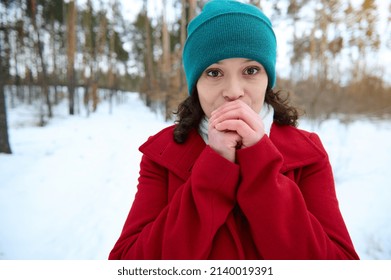 Cloe-up portrait of a middle aged woman in a red woolen coat and green hat warming her crossed hands because of cold weather standing in a snowy pine forest. Winter holidays, trip and travel concept