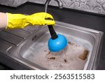 Clogged drain in kitchen sink. Hand in yellow gloves cleaning sink with plunger. Sink full of dirty water. Drain cleaning