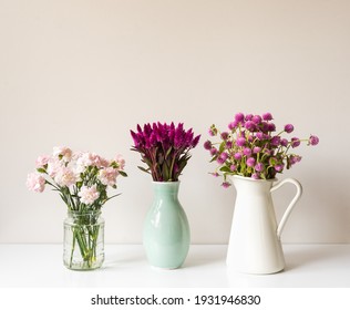 Cloose up of vases with pink and purple flowers incuding carnations, feathers, globe amaranths, on white table