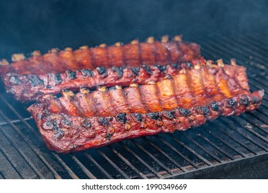 Close Up Of 2 Racks Of Bbq Ribs With Barbeque Sauce Cooking On Smoker Grill Plates Surrounded By Visible Smoke From The Smoker