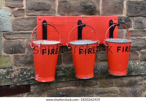 close up of 3 red Fire buckets hanging\
from hooks on a wooden plaque on a stone wall\
outside