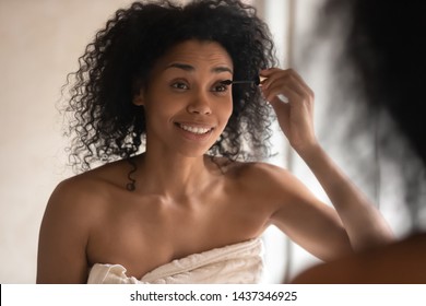 Close Up 30s African Woman Portrait, Female Wrapped In Towel After Shower And Morning Routine Look In Mirror Hold Liquid Tube Applicator Applying Make Up Mascara Make Her Lashes More Thicken Lengthen