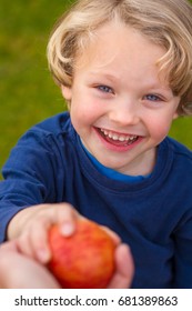 Close Up Of 5 Year Old Child With Blonde Hair And Blue Eyes Reaching Up Towards The Camera To Grab An Apple Being Offered To Him