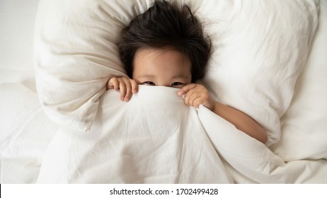 Close Up Above Top View Little Vietnamese Toddler Playful Girl Woke Up In The Morning Lying In Bed On Soft Pillow Comfortable Mattress Playing Looking At Camera Hides Her Face Under White Warm Blanket