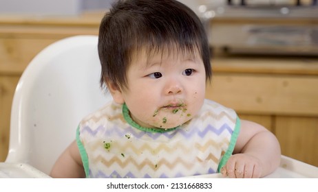 Close Up Adorable Female Toddler Wearing A Bib Is Getting Messy On Her Face While Being Fed With Solid Food From Spoon In Teh Dining Room At Home.