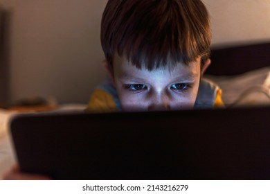 Close up of adorable kid laying down on bed by holding a smartphone at night. Little boy watching a movie on tablet, indoor. Shot of a cute little boy using a tablet while lying on the living room