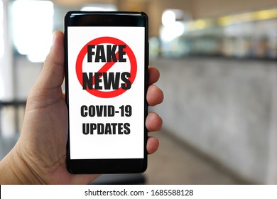 Close Up Adult Hand Holding A Smartphone With Stop Fake News Of Covid-19 Updates Text And Symbol On Display Screen, Blurred Cafe Background.