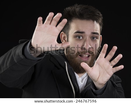 Close up Afraid Goatee Young Man in Dark Jacket, Raising his Hands Up While Looking at the Camera. Isolated on Black