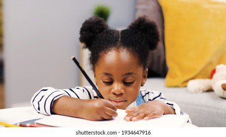 Close up African American cute little girl kid sitting in room in house drawing paper  writing in notebook doing homework  adorable child studying at home  leisure time  childhood concept