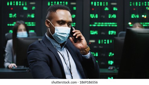 Close Up Of African American Handsome IT Programer Working On Desktop Computers In Data Center Control Room And Speaking On Smartphone. Young Security Analytic In Mask At Work Calling On Cellphone