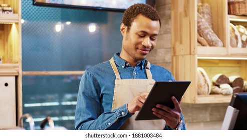 Close up of African American happy young handsome man worker in apron stands in bakery shop, texting and typing on tablet. Male seller browsing online on device. Business concept. Retail industry
