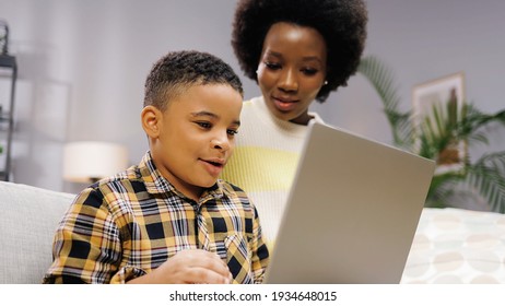 Close up of African American joyful young small cute son browsing online on laptop sitting on sofa with beautiful mother. Little boy with mom studying on computer surfing internet, childhood concept - Powered by Shutterstock