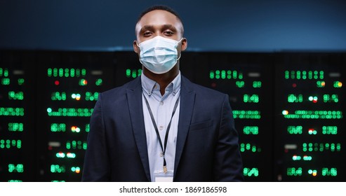 Close up of African American male IT administrator wearing medical mask standing in modern data center with many processors. Portrait of handsome young man security engineer in suit. Business concept