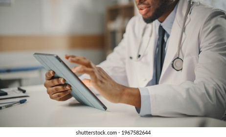Close Up of African American Male Doctor Wearing White Coat Working on Tablet Computer at His Office. Medical Health Care Professional Working with Test Results, Patient Treatment Planning. - Powered by Shutterstock