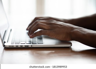 Close up of African American male employee busy typing texting on keyboard on computer, focused biracial man browsing Internet working on modern laptop on wooden desk indoors