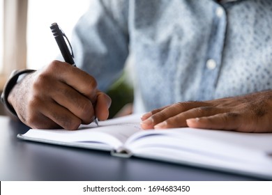 Close up of african American male student busy handwriting in notebook preparing for exam or test, biracial man worker make notes or list writing down, planning or thinking at workplace in office
