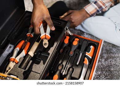 Close up of african american man in casual clothes taking pliers out of box at home. Focus on plastic black container with variety of modern tools for housework.