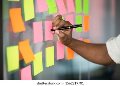 Close up african american manager writing notes on colorful sticky notes on kanban board. Mixed race employee managing project workflow, scheduling tasks or planning strategy on glass wall at office. - Shutterstock ID 1562219701