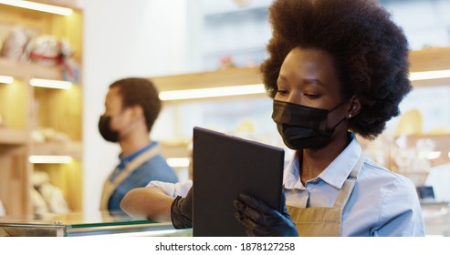 Close Up Of African American Pretty Woman Seller In Black Mask And Gloves Standing In Coffee Shop Typing On Tablet At Workplace In Bakery Shop While Her Male Co-worker Working On Background In Bakery