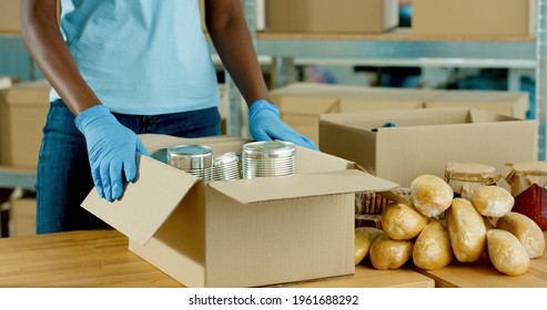 Close Up Of African American Social Worker Hands In Protective Gloves Packing Food Products In Donation Box Working In Charity Organization. Food Donations, Coronavirus. Donations And Volunteer