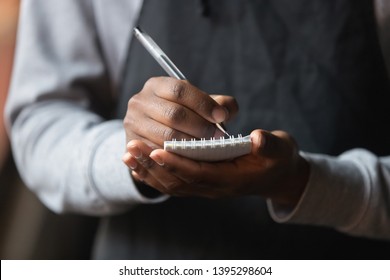 Close Up African American Waiter Hands With Notebook, Taking Customer Order In Cafe Or Restaurant, Coffeehouse Worker Wearing Black Apron Serving Client, Writing Down, Service Concept