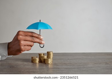 Close up of african american woman hand holding blue umbrella over stack of coins on table. Hands of financial advisor saving money. Businesswoman protect saving and money with copy space. - Powered by Shutterstock