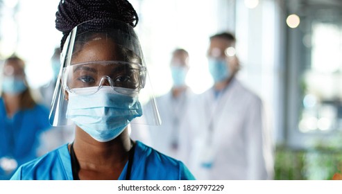 Close Up Of African American Woman Physician In Face Shield Looking To Camera In Clinic. Portrait Of Female Doctor Or Nurse In Medical Mask And Goggles In Hospital. Docs Talking On Background Covid-19