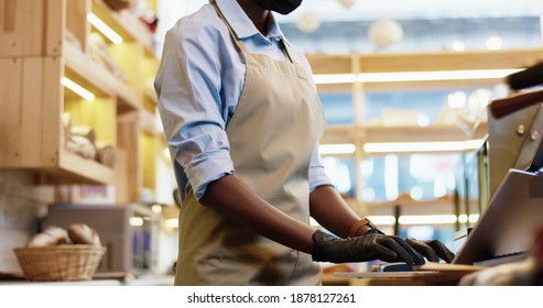 Close Up Of African American Woman Seller In Apron In Black Mask And Gloves Typing On Laptop Standing At Workplace In Bakery Shop While Her Male Co-worker Working On Background. Sales Concept