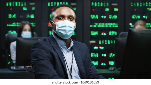 Close Up Of African American IT Worker In Mask Working On Desktop Computers In Data Center Control Room And Looking At Camera Male Security Engineer Working On Computer. Female Analytics On Background