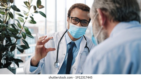 Close Up Of African American Young Handsome Male Doctor Specialist In Medical Mask Talking With Caucasian Old Patient Explaining New Coronavirus Vaccine On Medical Consultation, Vaccination Concept