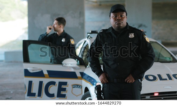 Close up african american young man cops stand near\
patrol car look around background his colleague enforcement happy\
officer police uniform auto safety security communication control\
policeman close