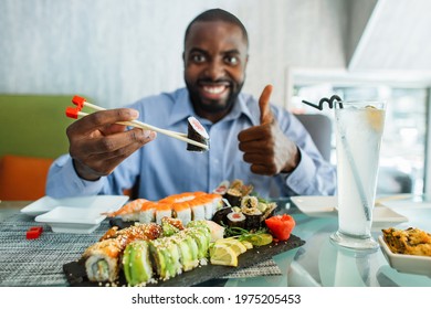 Close Up Of African Man Eating Sushi At Cafe. Handsome Smiling Black Man Eating Sushi Rolls, Using Chopsticks, Doing Ok Excellent Sign, Thumb Up With Fingers. Focus On His Hands With Roll.