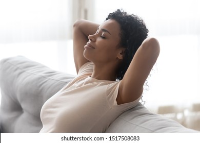 Close Up African Serene Woman Closed Eyes Putting Hands Behind Head Resting Leaned On Comfortable Couch Enjoy Fresh Cool Air At Hot Summer Day In Living Room At Home, No Stress Lazy Weekend Concept