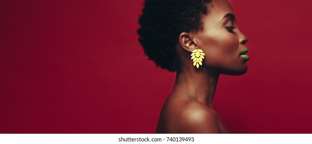Close up of african woman wearing ear rings and vivid makeup against red background. Profile view of female model with beautiful skin.