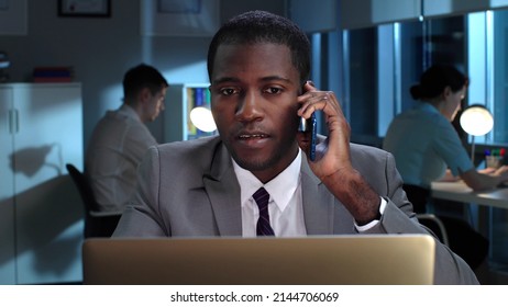 Close Up Of African-American Lawyer Having Late Phone Call With Client. Businessman Sit At Desk With Laptop And Talk On Smartphone In Dark Office