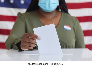 Close up of African-American woman wearing mask putting vote bulletin in ballot box and looking at camera while standing against American flag on election day, copy space - Shutterstock ID 1810359352