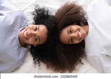 Close up afro teen daughter lying on bed with mother smile look at camera, top above view. Sisters with curly hairs relaxing during sleepover have lazy weekends natural beauty candid portrait concept