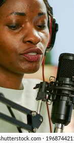 Close up of afro woman vlogger making new video blog in home studio talking in professional microphone with audience. Speaking during livestreaming, blogger discussing in podcast wearing headphones.