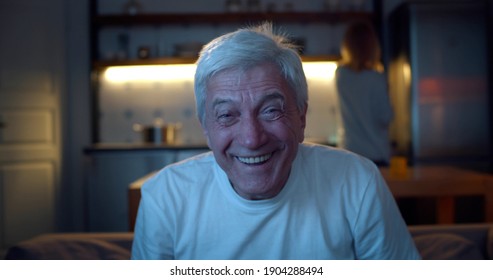 Close Up Of Aged Man Watching Tv In Evening And Laughing Relaxing On Couch. Happy Senior Male Watching Comedy Show In Living Room With Wife Cooking In Kitchen On Background